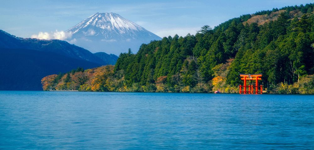 A serene blue lake stretches toward a forested shoreline with a red torii gate standing near the water’s edge. Snow-capped mountains rise in the background under a clear blue sky.