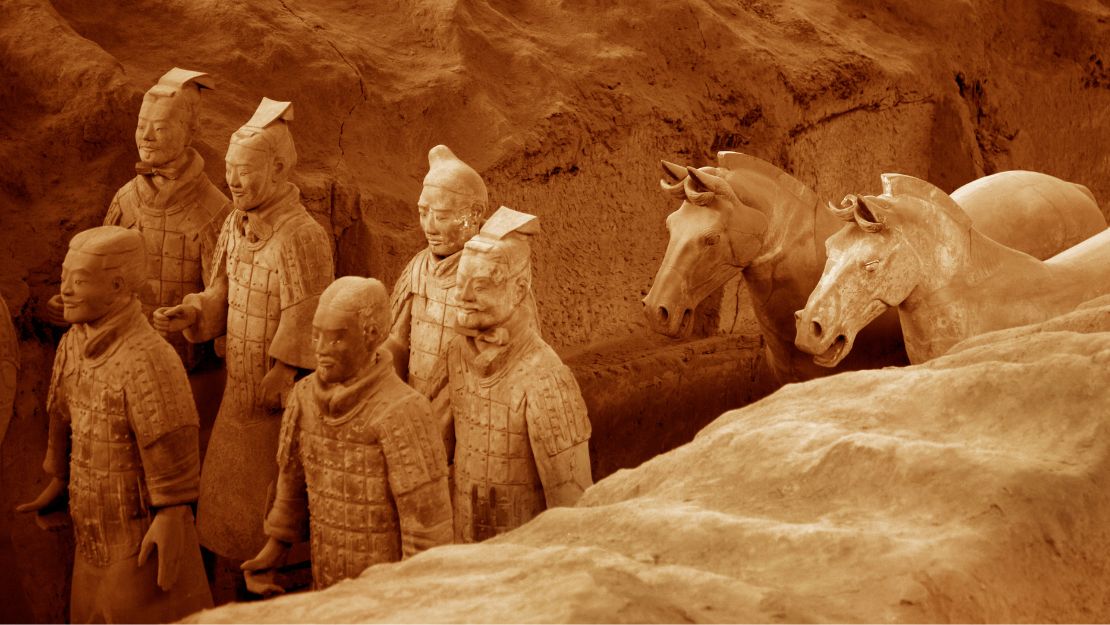 Ancient terracotta warrior statues stand in rows inside an excavation site. The earthy tones of the figures and surrounding soil highlight their historical significance.