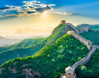 The Great Wall of China winds across lush green mountains under a bright sky. Its stone structure stretches into the distance, showcasing the scale and grandeur of this historic landmark.