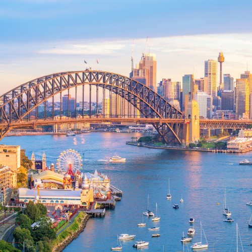 Scenic view of Sydney Harbour at sunset, featuring the iconic Harbour Bridge, skyline with skyscrapers, and boats on the sparkling water below.