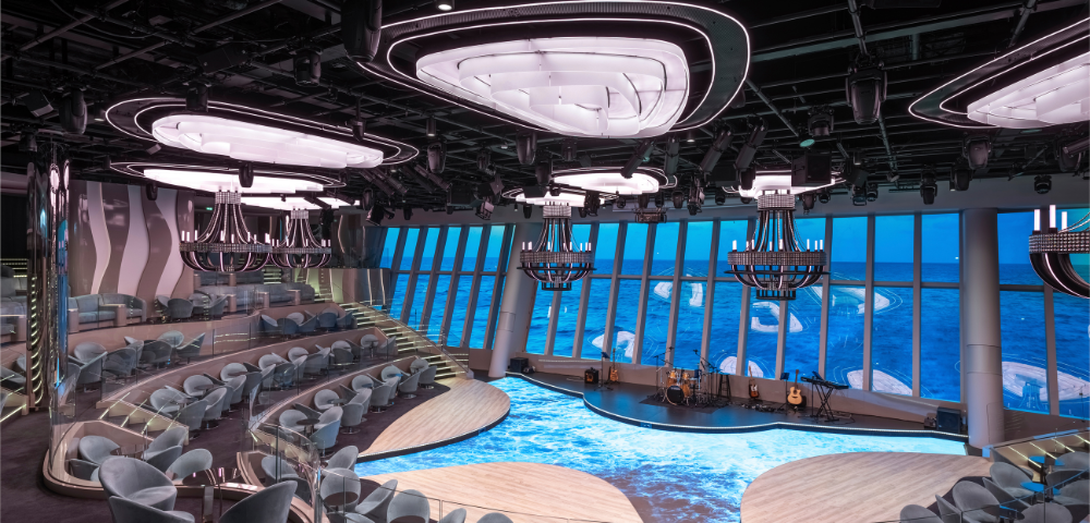 A modern performance space with tiered seating, large windows showcasing ocean views, and stylish chandeliers, illuminated in soft lighting.