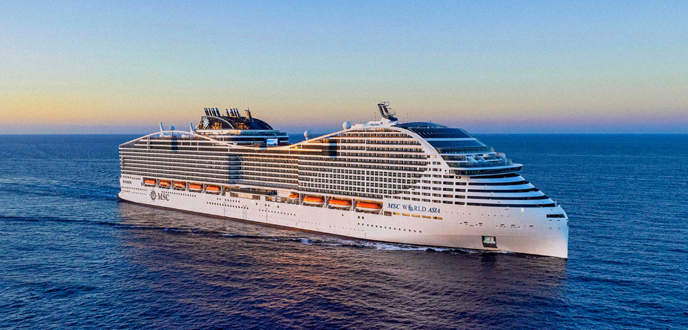 A large, modern cruise ship sails on calm open water under a clear sky at sunset.
