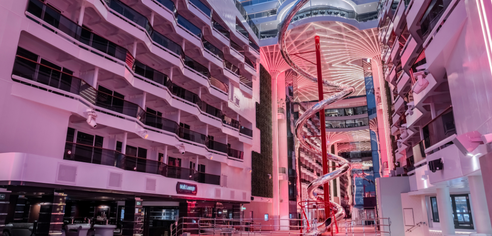 A large open atrium with balconies, a central spiral slide, and pink lighting, possibly inside a modern cruise ship or hotel.