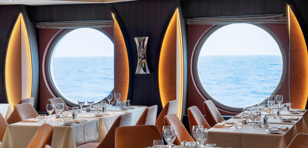 A cozy dining room featuring tables and chairs, with large windows offering a view of the ocean. The setup includes tableware such as wine glasses and tablecloths, creating an inviting atmosphere for indoor dining.