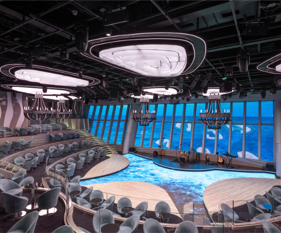 A modern performance space with tiered seating, large windows showcasing ocean views, and stylish chandeliers, illuminated in soft lighting.