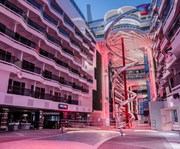 A large open atrium with balconies, a central spiral slide, and pink lighting, possibly inside a modern cruise ship or hotel.
