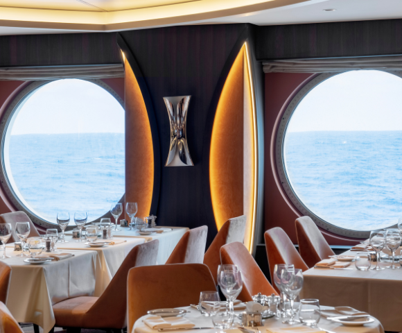 A cozy dining room featuring tables and chairs, with large windows offering a view of the ocean. The setup includes tableware such as wine glasses and tablecloths, creating an inviting atmosphere for indoor dining.