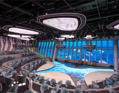 A modern performance space with tiered seating, large windows showcasing ocean views, and stylish chandeliers, illuminated in soft lighting.