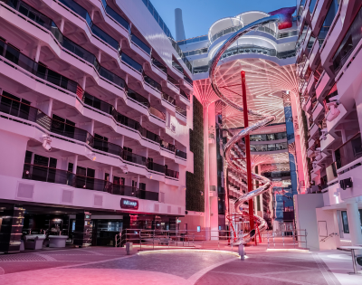 A large open atrium with balconies, a central spiral slide, and pink lighting, possibly inside a modern cruise ship or hotel.