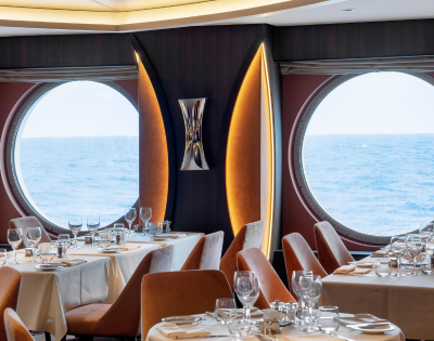 A cozy dining room featuring tables and chairs, with large windows offering a view of the ocean. The setup includes tableware such as wine glasses and tablecloths, creating an inviting atmosphere for indoor dining.