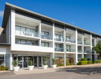 Modern three-story building with a sleek facade, large windows, and balconies. The entrance is flanked by potted plants. Bright, sunny day.