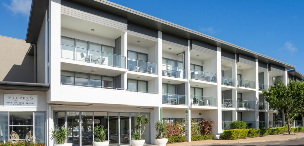 Modern three-story building with a sleek facade, large windows, and balconies. The entrance is flanked by potted plants. Bright, sunny day.