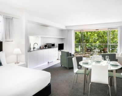 Modern open-plan living space with white decor, featuring a kitchen, dining area, and lounge. Large glass doors open to a sunny patio with a marina view.