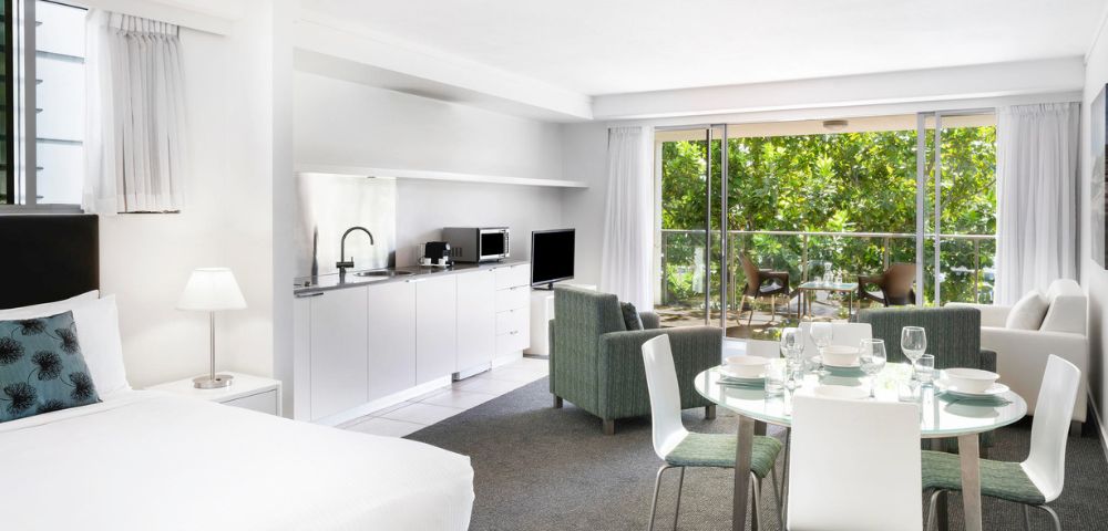 Modern open-plan living space with white decor, featuring a kitchen, dining area, and lounge. Large glass doors open to a sunny patio with a marina view.