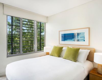 A bright bedroom with a white bed, green pillows, and a framed abstract artwork. Large window shows trees outside, creating a serene atmosphere.