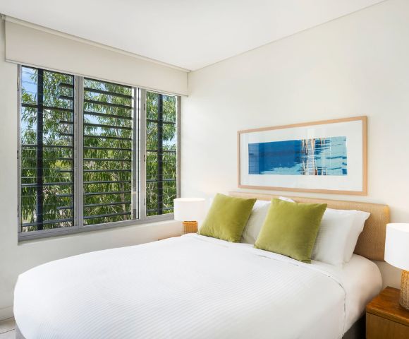 A bright bedroom with a white bed, green pillows, and a framed abstract artwork. Large window shows trees outside, creating a serene atmosphere.
