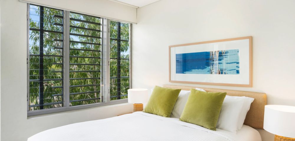 A bright bedroom with a white bed, green pillows, and a framed abstract artwork. Large window shows trees outside, creating a serene atmosphere.