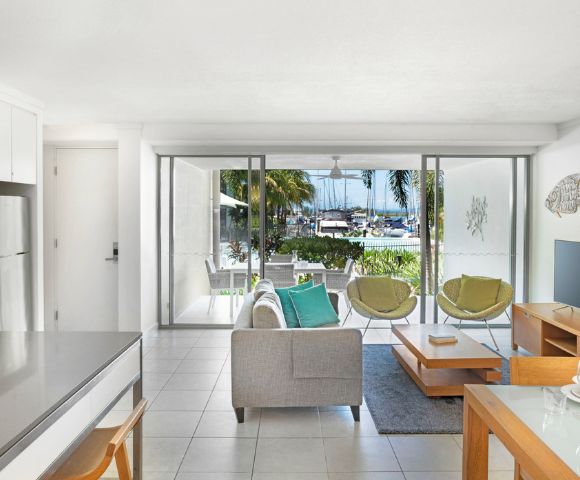 Modern living room with neutral tones, featuring a sofa, two green chairs, a TV, and a coffee table. Large glass doors open to a marina view. Relaxed ambiance.