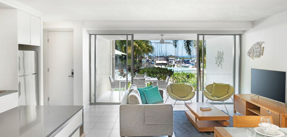 Modern living room with neutral tones, featuring a sofa, two green chairs, a TV, and a coffee table. Large glass doors open to a marina view. Relaxed ambiance.