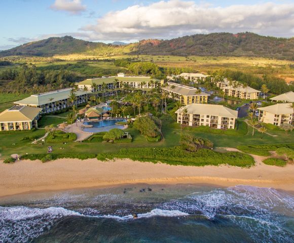 Aerial view of a beachfront resort with multiple buildings, a central pool area, and lush greenery. The ocean waves gently lap against the sandy shore, evoking a tranquil, vacation-like atmosphere. Hills and fields stretch in the background under a partly cloudy sky.