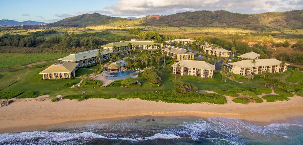 Aerial view of a beachfront resort with multiple buildings, a central pool area, and lush greenery. The ocean waves gently lap against the sandy shore, evoking a tranquil, vacation-like atmosphere. Hills and fields stretch in the background under a partly cloudy sky.