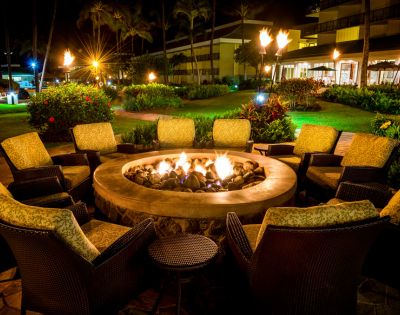 Cozy outdoor setting with wicker chairs and cushions encircling a stone fire pit, glowing warmly at night. Lush garden and lit lamps create a serene atmosphere.