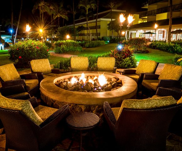 Cozy outdoor setting with wicker chairs and cushions encircling a stone fire pit, glowing warmly at night. Lush garden and lit lamps create a serene atmosphere.