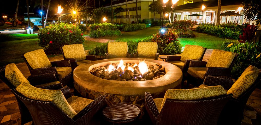 Cozy outdoor setting with wicker chairs and cushions encircling a stone fire pit, glowing warmly at night. Lush garden and lit lamps create a serene atmosphere.