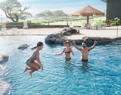 A young girl is jumping into a pool, splashing water. Two adults, a man and a woman, smile and cheer her on. The scene is joyful and sunny.