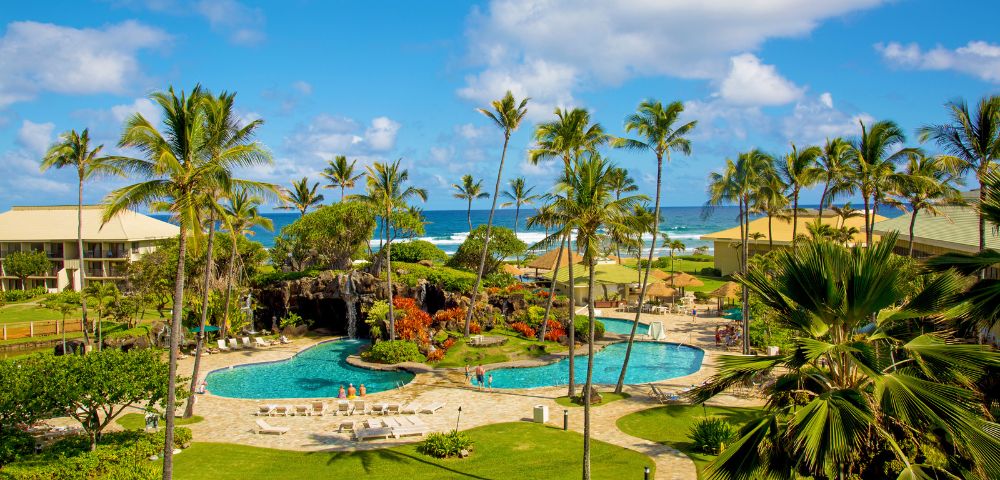 Tropical resort scene with palm trees, blue pools, and ocean view under a bright sky. Lush gardens and sun loungers create a relaxing atmosphere.