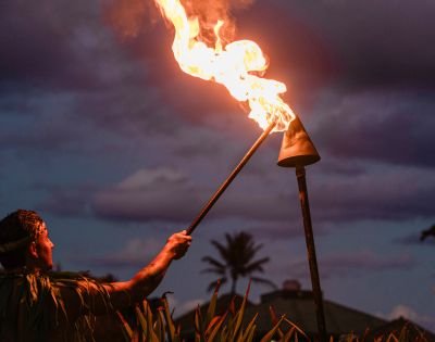 A person in traditional attire lights a tiki torch at sunset, with dramatic clouds and a silhouette of a palm tree in the background. The scene evokes a warm, tropical ambiance.