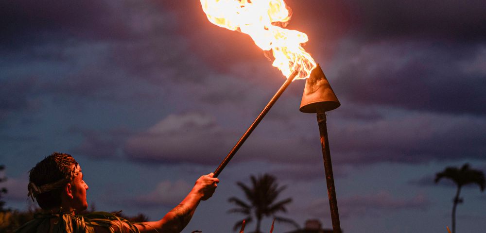 A person in traditional attire lights a tiki torch at sunset, with dramatic clouds and a silhouette of a palm tree in the background. The scene evokes a warm, tropical ambiance.