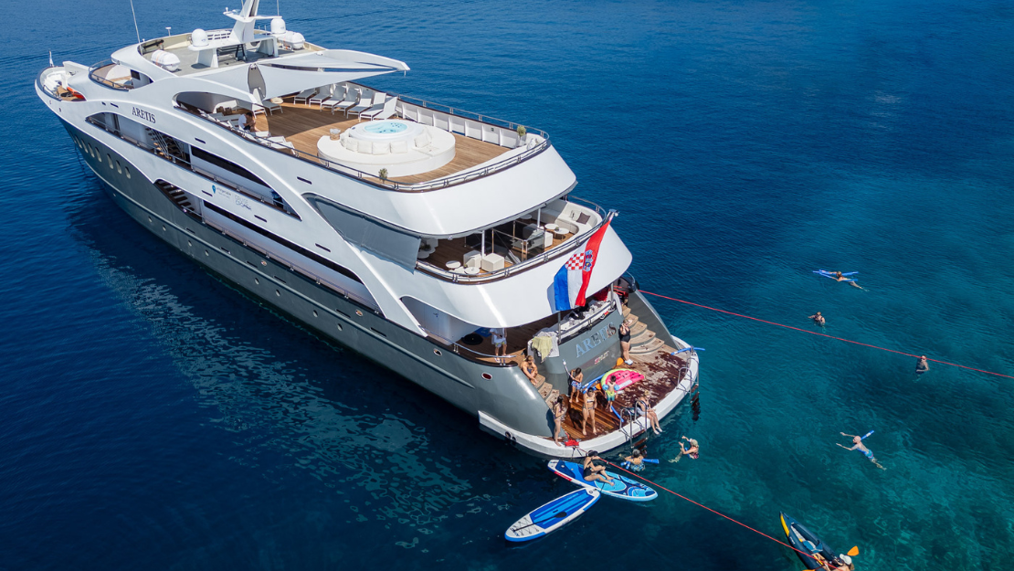 A luxurious yacht anchored in clear blue water, with people swimming, paddleboarding, and enjoying the sun on the deck.