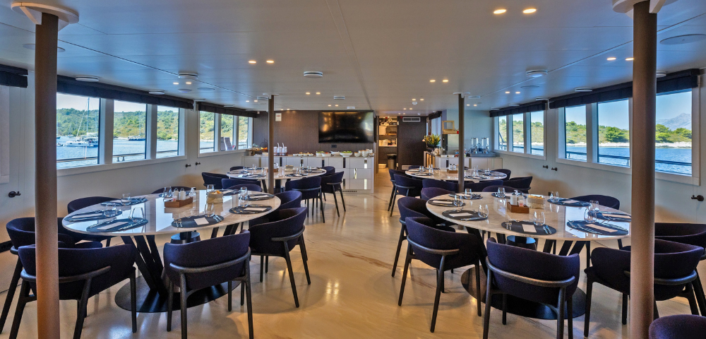 A room featuring multiple tables and chairs arranged for dining, with a ceiling overhead and a floor underneath. The space is designed for social gatherings, resembling a cafeteria or restaurant setting.