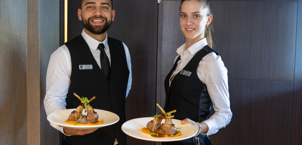 A man and woman standing indoors, smiling while holding plates of food. The background features a wall, and they are both dressed in casual clothing.