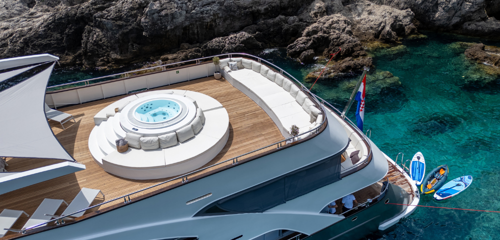 A luxury yacht with a spacious deck, featuring a hot tub, lounge seating, and jet skis beside clear blue waters and rocky surroundings.
