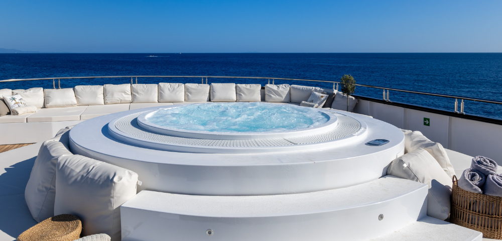 A luxurious hot tub on a sunlit deck, surrounded by plush white seating, overlooks a serene blue ocean under a clear sky.