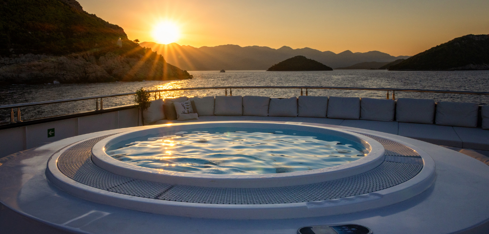 A luxurious hot tub on a sunlit deck, surrounded by plush white seating, overlooks a serene blue ocean under a clear sky.