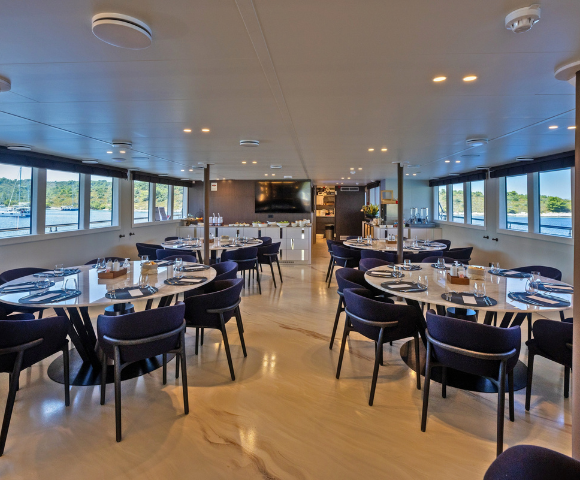 A room featuring multiple tables and chairs arranged for dining, with a ceiling overhead and a floor underneath. The space is designed for social gatherings, resembling a cafeteria or restaurant setting.