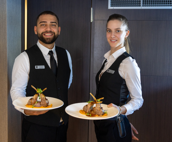 A man and woman standing indoors, smiling while holding plates of food. The background features a wall, and they are both dressed in casual clothing.