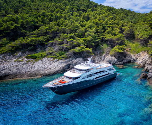 A luxurious yacht anchored in crystal-clear turquoise waters near a rocky, green-covered coastline.