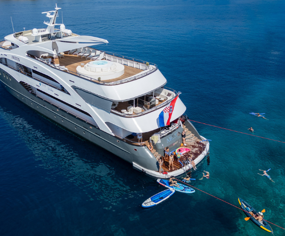 A luxurious yacht anchored in clear blue water, with people swimming, paddleboarding, and enjoying the sun on the deck.