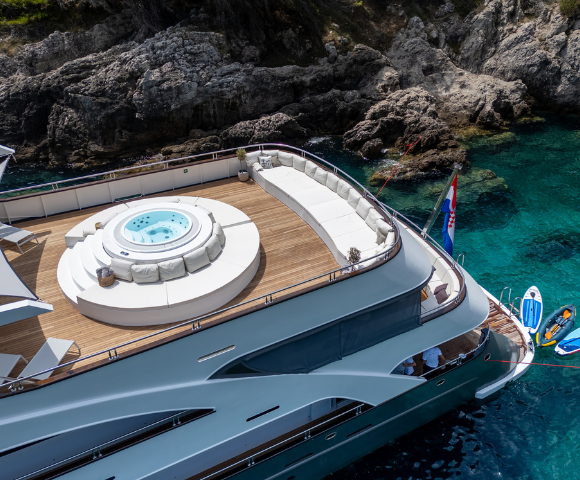 A luxury yacht with a spacious deck, featuring a hot tub, lounge seating, and jet skis beside clear blue waters and rocky surroundings.