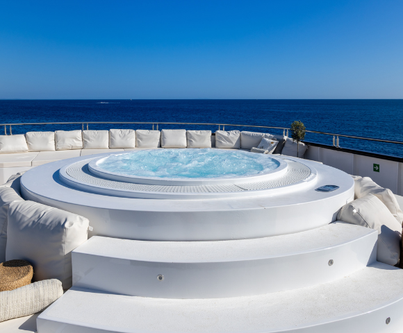A luxurious hot tub on a sunlit deck, surrounded by plush white seating, overlooks a serene blue ocean under a clear sky.