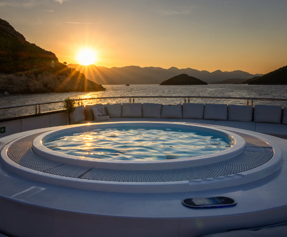 A luxurious hot tub on a sunlit deck, surrounded by plush white seating, overlooks a serene blue ocean under a clear sky.
