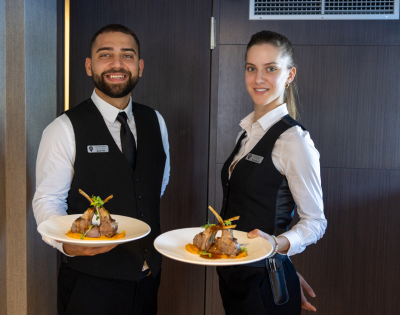 A man and woman standing indoors, smiling while holding plates of food. The background features a wall, and they are both dressed in casual clothing.