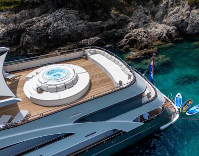 A luxury yacht with a spacious deck, featuring a hot tub, lounge seating, and jet skis beside clear blue waters and rocky surroundings.
