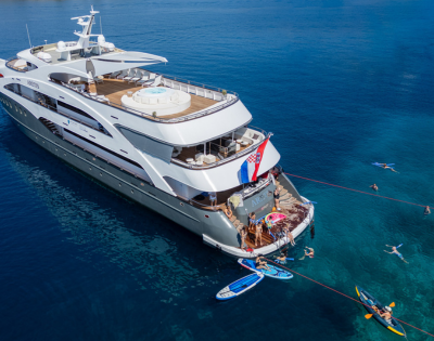 A luxurious yacht anchored in clear blue water, with people swimming, paddleboarding, and enjoying the sun on the deck.