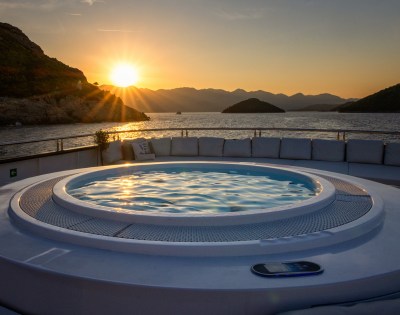 A luxurious hot tub on a sunlit deck, surrounded by plush white seating, overlooks a serene blue ocean under a clear sky.