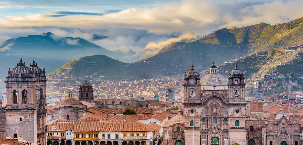 Historic cityscape of Cusco, Peru, with ornate cathedral towers and colonial buildings in the foreground. Misty mountains rise under a blue sky.